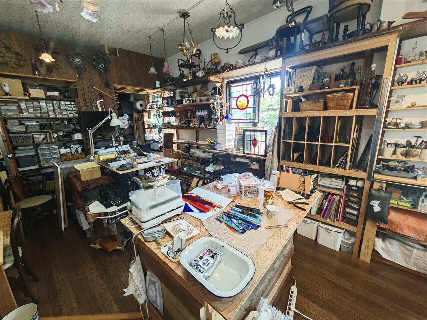 京都・大原野の工房の全景。中央の作業台にガラスと道具が広がり、壁一面に工具と部材の棚、天井からアンティークの照明が下がる。奥の窓には制作したステンドグラスが納まっている