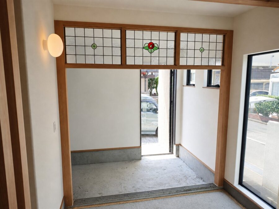 玄関ホールの欄間に納めた3枚組のステンドグラス。木枠の格子に質感の異なる型板ガラスを並べ、中央のパネルに赤いバラと緑の葉、両側のパネル中央に緑のロンデルガラスを配している