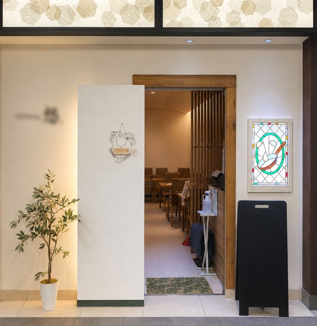 改装後の洋食店エントランスを正面から見た様子。白い壁と無垢材の枠に替え、左にオリーブの鉢、右の壁に背面から照らしたステンドグラスを掛けている