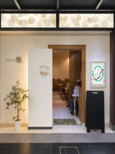 改装後の洋食店エントランスを正面から見た様子。白い壁と無垢材の枠に替え、左にオリーブの鉢、右の壁に背面から照らしたステンドグラスを掛けている