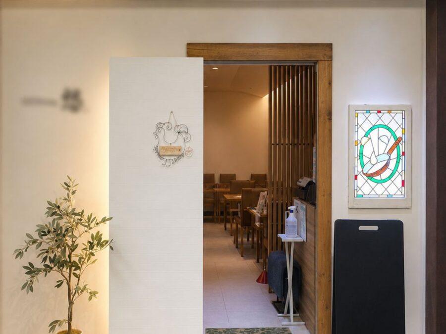 改装後の洋食店エントランスを正面から見た様子。白い壁と無垢材の枠に替え、左にオリーブの鉢、右の壁に背面から照らしたステンドグラスを掛けている