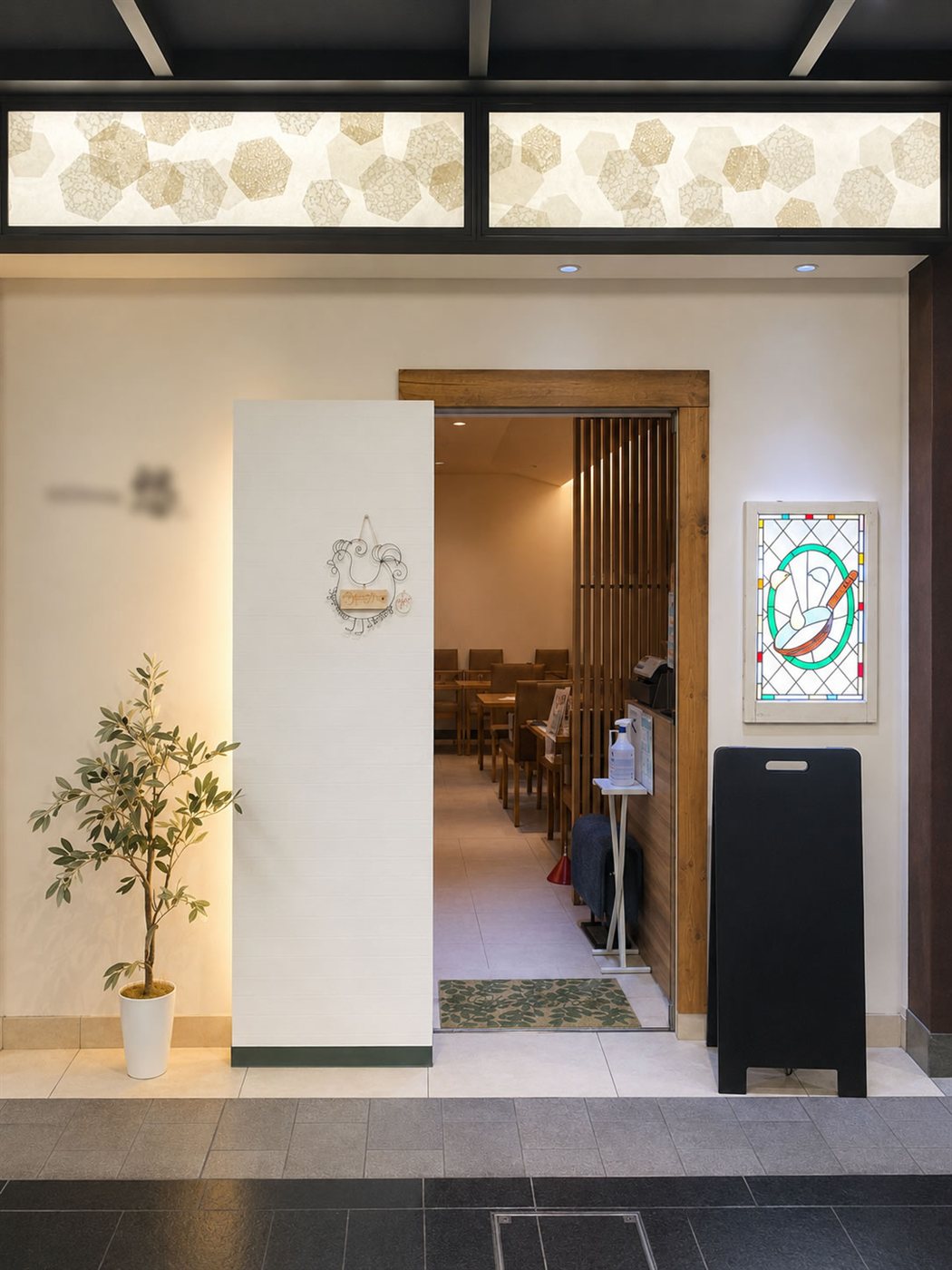 改装後の洋食店エントランスを正面から見た様子。白い壁と無垢材の枠に替え、左にオリーブの鉢、右の壁に背面から照らしたステンドグラスを掛けている